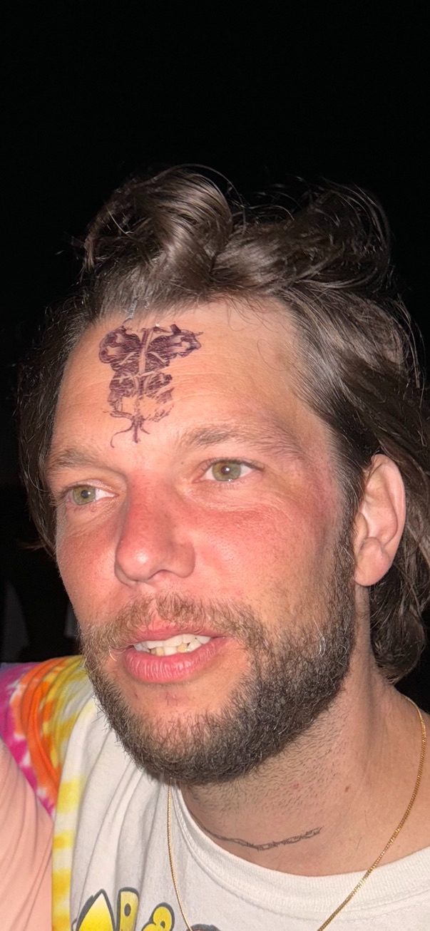 A man with a temporary caduceus tattoo on his forehead, photographed at night in a tie-dye shirt.