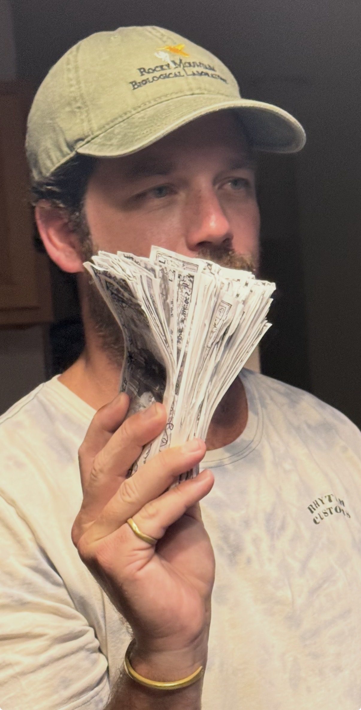 A man lifts a thick fan of currency notes beside his face, gold bracelet at his wrist, quietly satisfied.