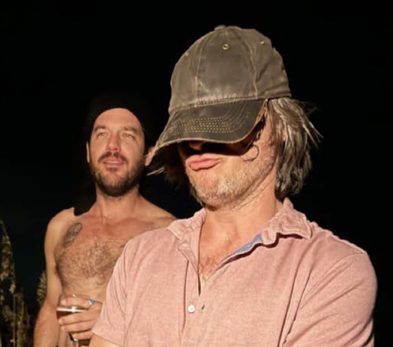 One man displays a temporary face tattoo while standing beside another man indoors.