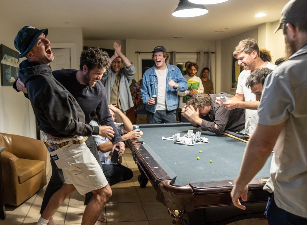 A dramatically lit scene of men frozen around a gaming table at the decisive moment of a dice hand, arms raised in shock.