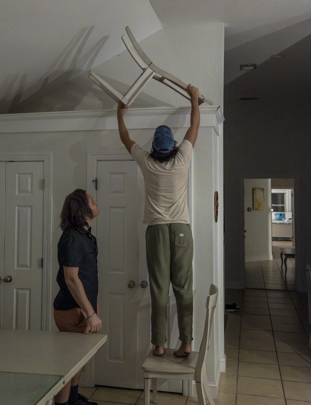 Late at night, one man watches another lift a chair onto the fireplace mantle.