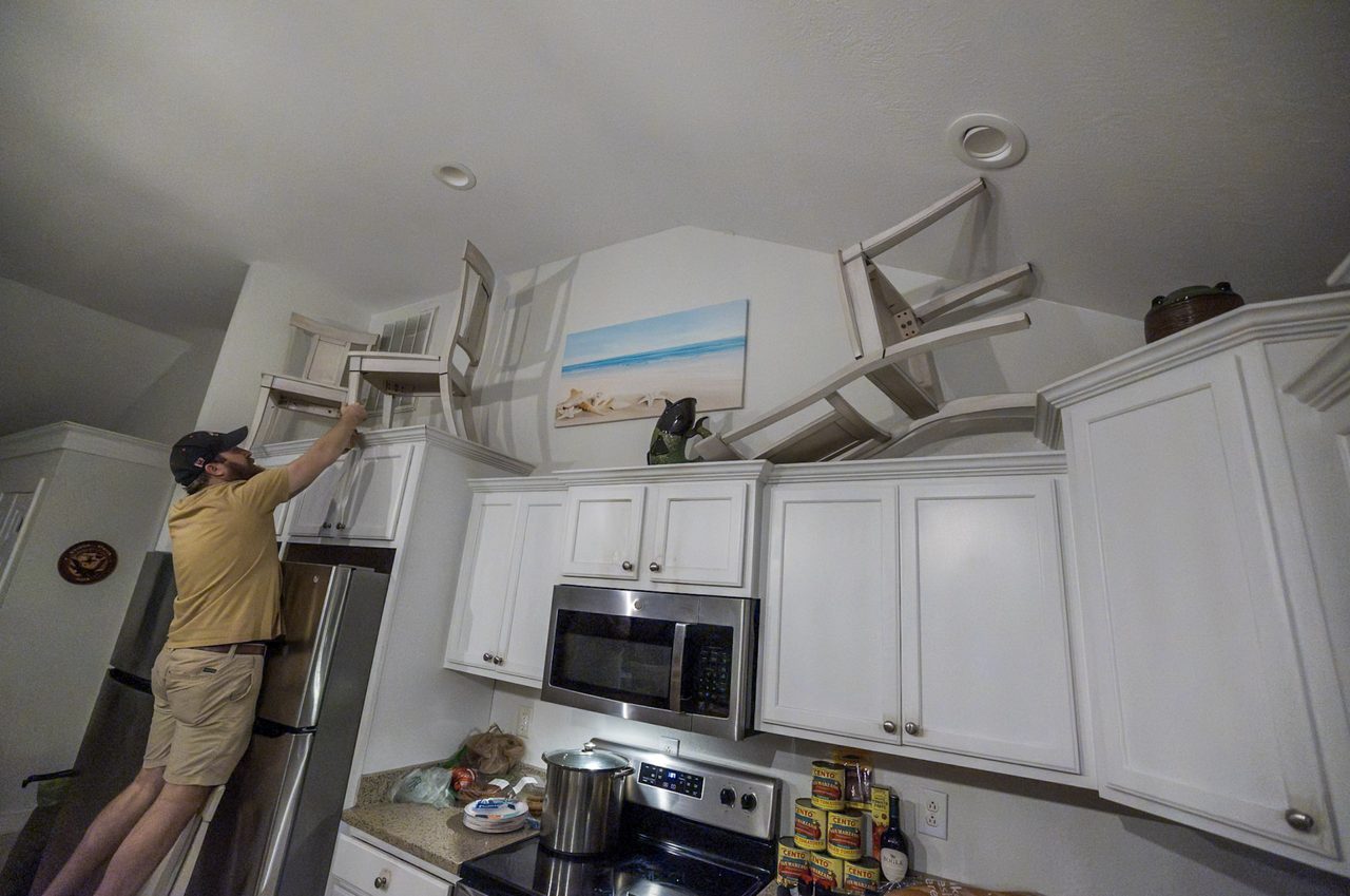 A man balances another chair on top of a growing stack of chairs on the fireplace mantle.
