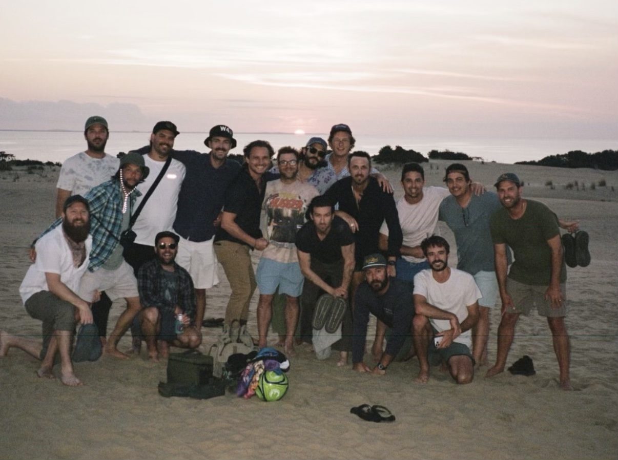The full group posed together on beach dunes at dusk, bags and boards on the sand, a pale sunset sky behind them.