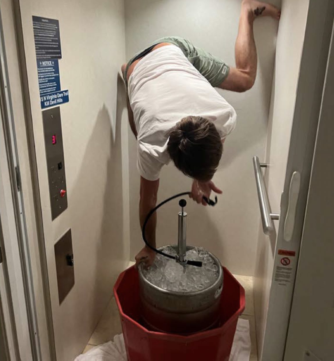 A man does a keg stand inside a residential elevator while others hold his legs.