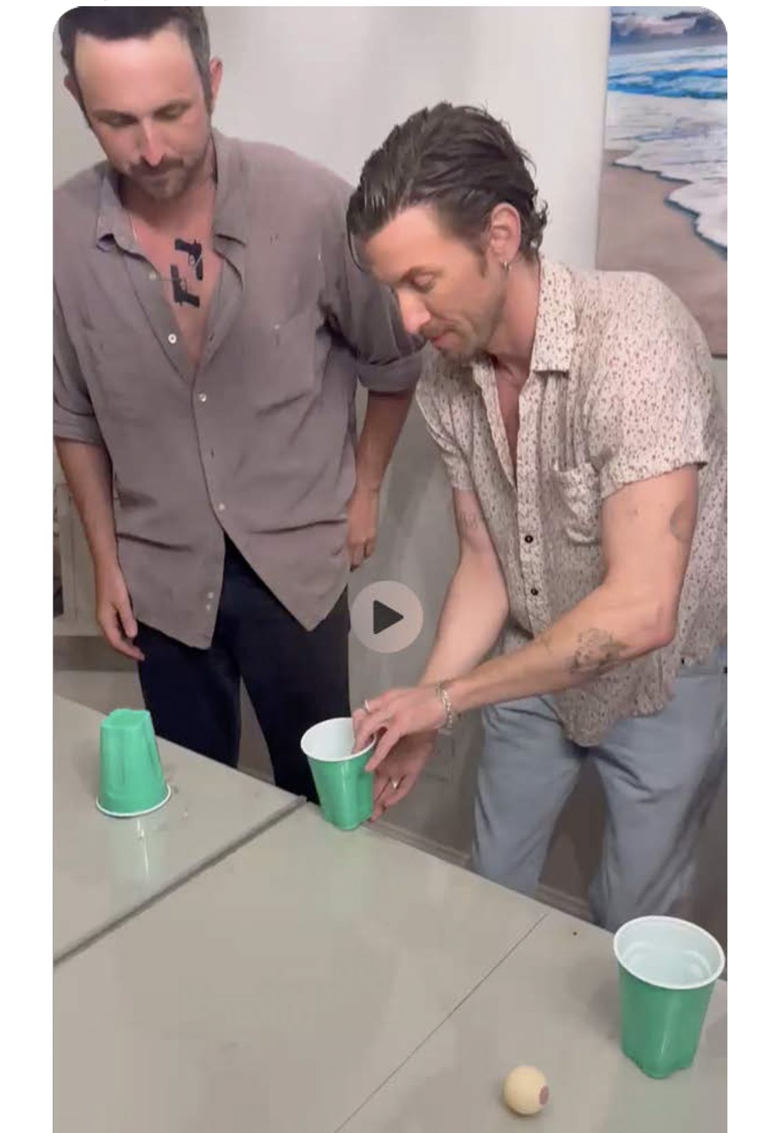 Two men mid-game at a table of cups, one flipping a cup at the table's edge.