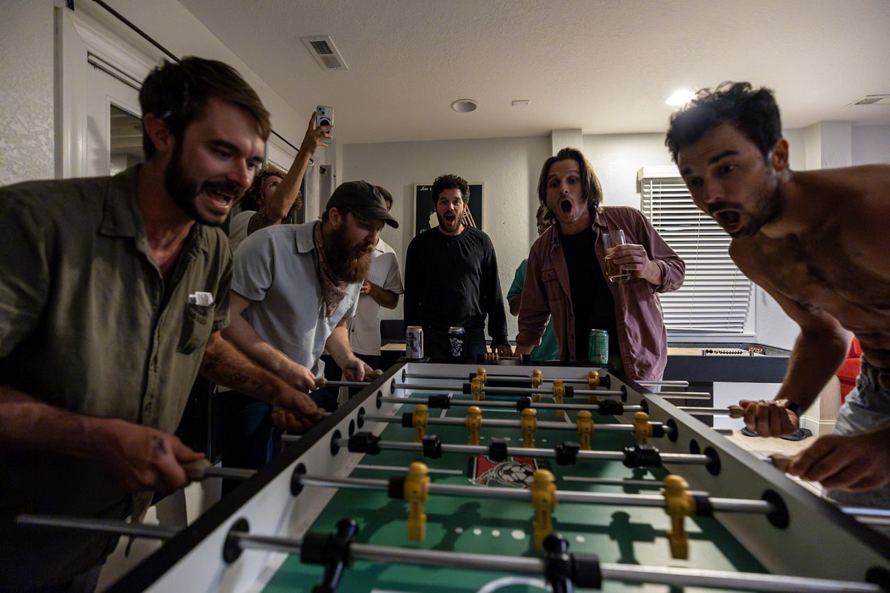 Four men crowd around a foosball table with paper notes stacked along its rail.