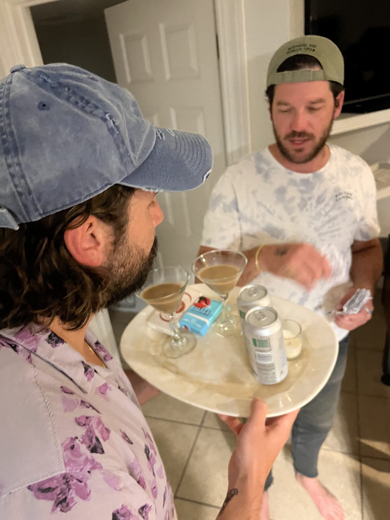 Two men exchange a drink and paper notes across a kitchen counter.