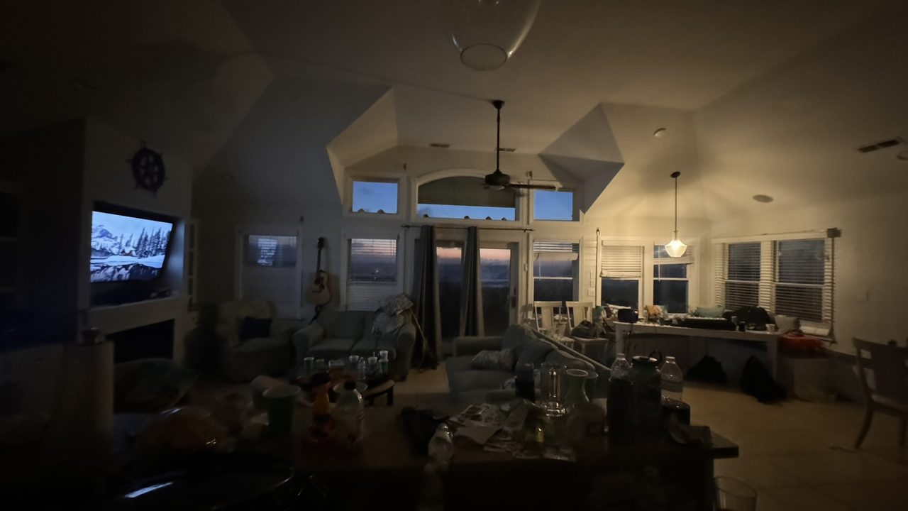 A dim living room at dawn strewn with bottles, cups, and gear, sunrise showing through the beach-house windows.
