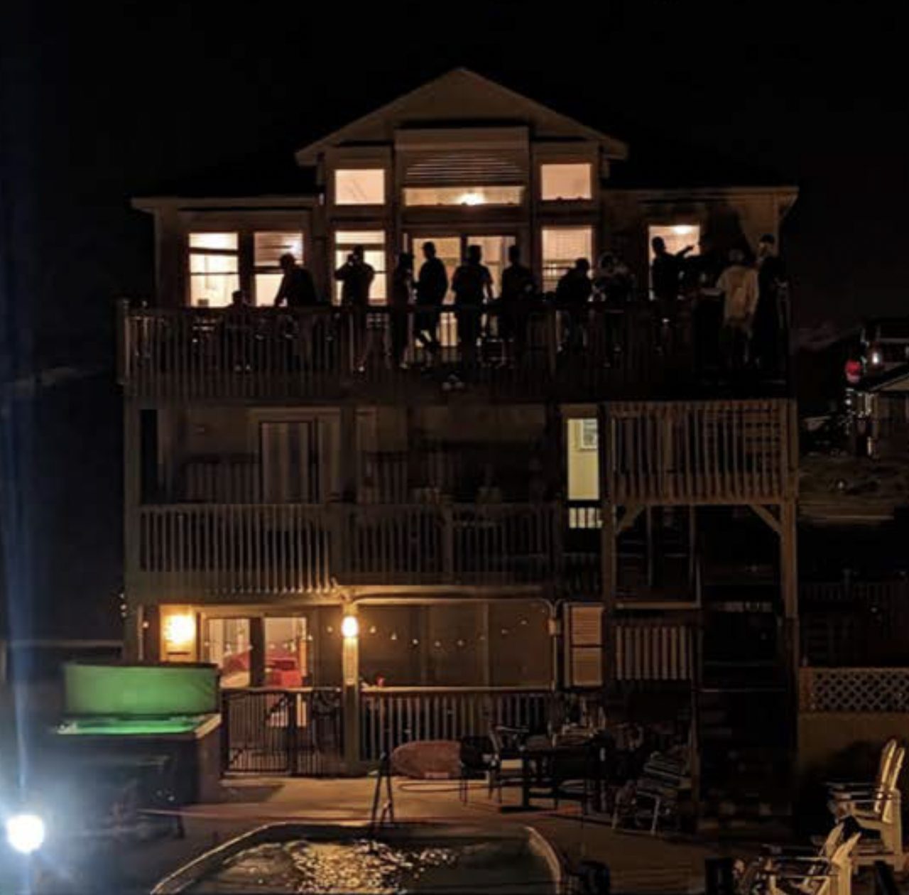 A three-story beach house at night with men gathered along its lit balconies.