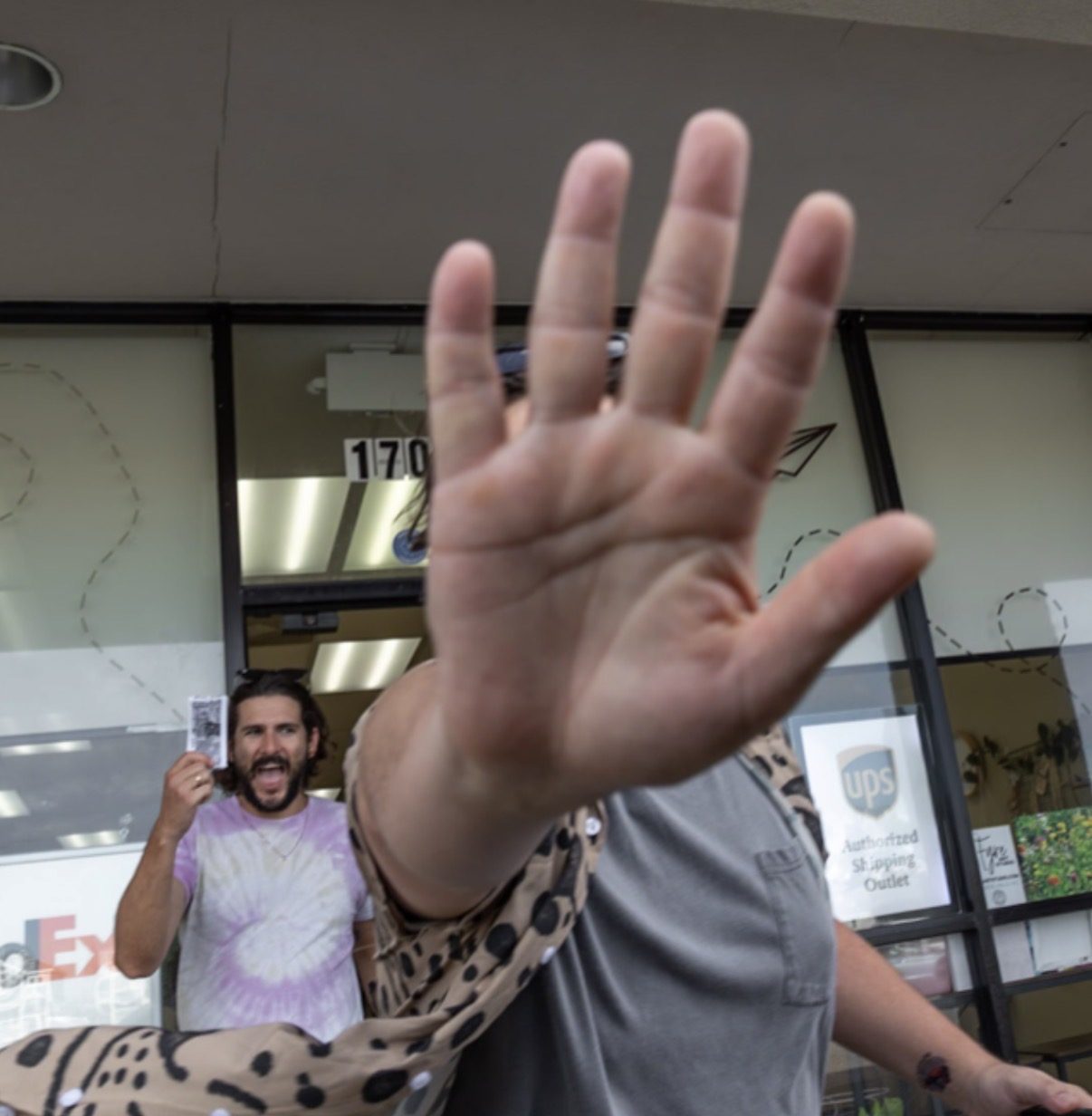 A hand thrust toward the camera lens, blocking the shot, while a man holding printed notes stands in the background outside a store.