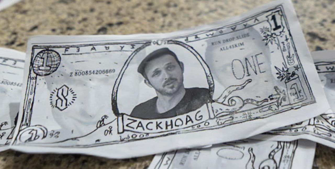 A single hand-drawn one-unit note with a portrait, ornate border, and the name ZACK HOAG, photographed on a granite countertop.
