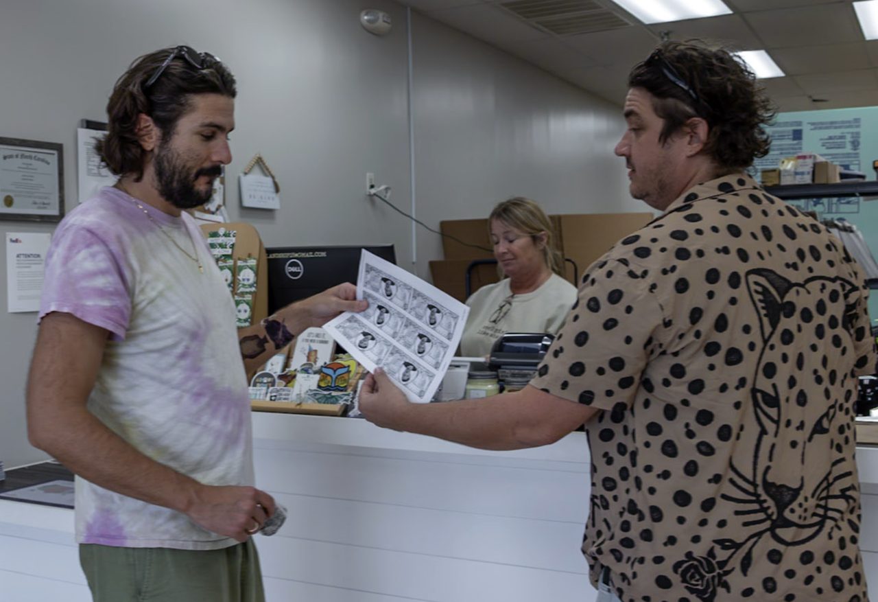 Two men inside a print shop hold freshly printed sheets of currency while a store employee stands behind the counter in the background.