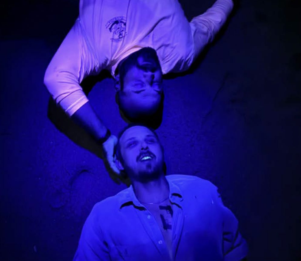 Two men on a dark beach lit by ultraviolet light, their clothes and skin glowing.